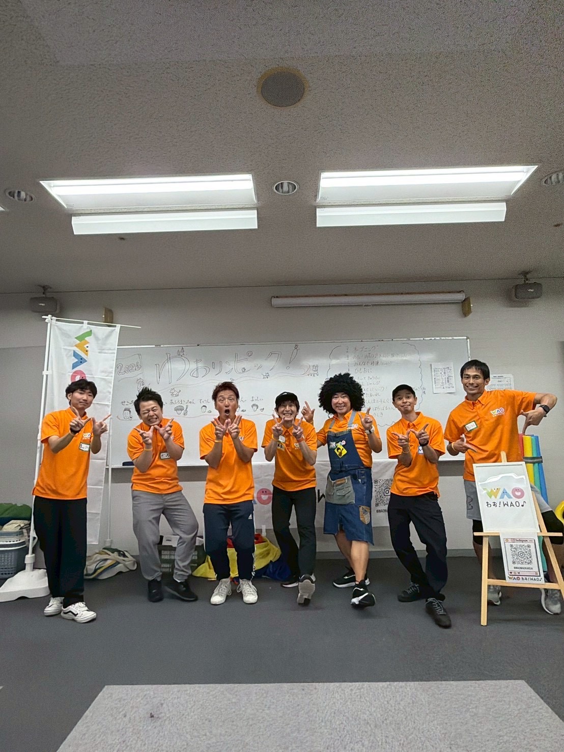 ホワイトボードの前でおそろいのオレンジのポロシャツを着てポーズをとる、わお！WAO♪のメンバー7人