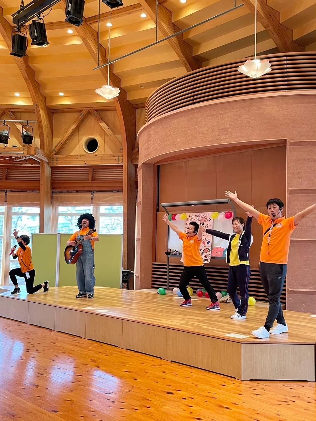 明るいホールのステージで子どもたちと一緒にパフォーマンスをするメンバー